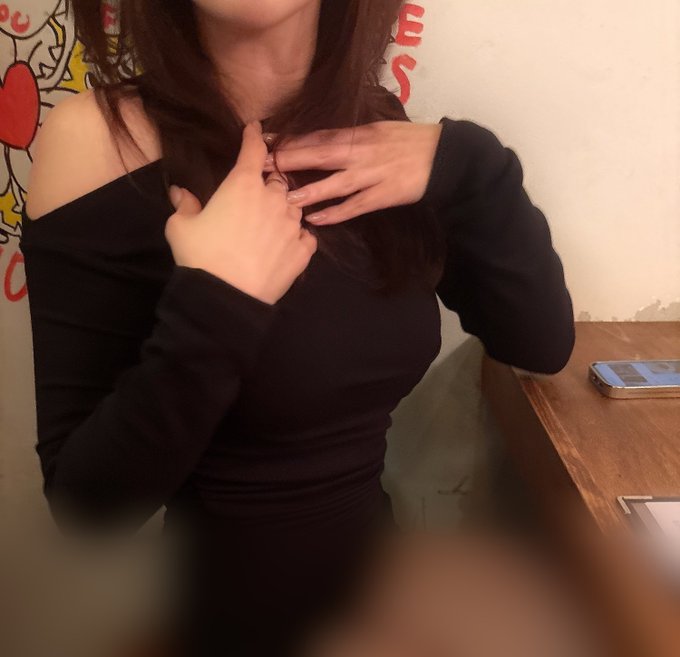 A person with long dark hair wearing a black off-shoulder top poses with hands near their chest in front of a wall with cartoon artwork.