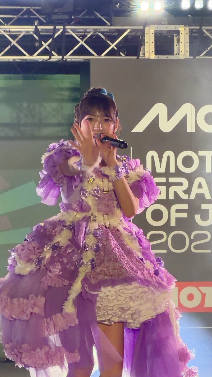 A young performer in an ornate purple ruffled gown sings into a microphone on stage, with partial text behind her reading "MO... GRA... OF J... 202..." suggesting a MotoGP Grand Prix of Japan 2023 event, connecting to the #MotoGP hashtag and the hashtag #塩川莉世 (Riyoshi Shiokawa) of the idol group #SWEETSTEADY performing the song "Sui Sui Skip."