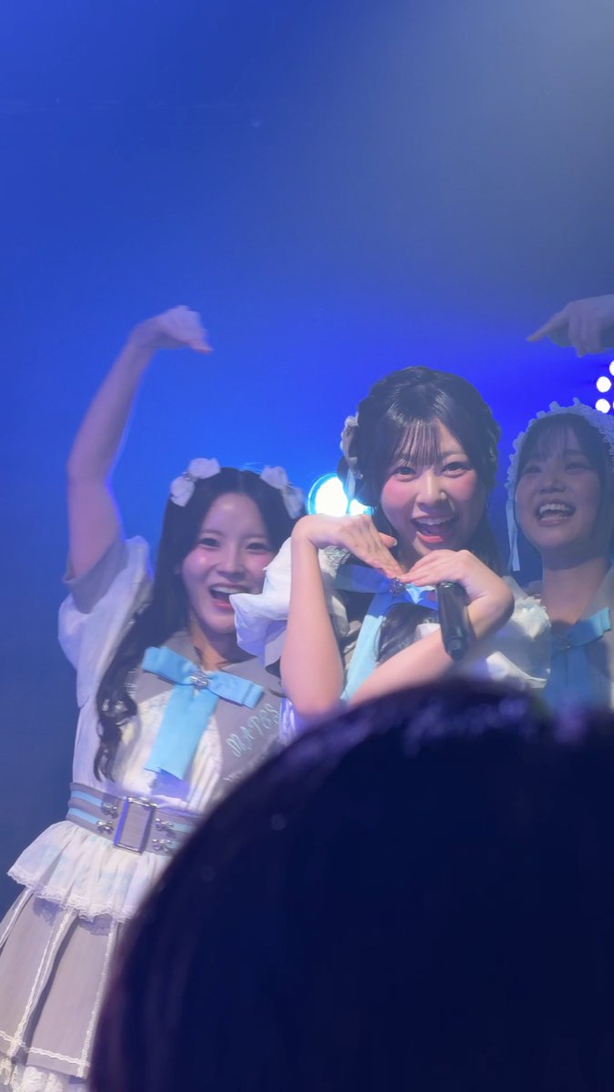 Three young women in matching white and blue idol dresses with bows pose energetically on a blue-lit stage, smiling and making heart shapes with their arms.