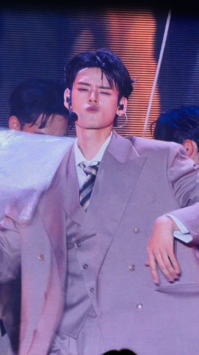A male performer on stage wearing a beige three-piece suit with a striped tie, eyes closed in a dramatic expression, with other dancers visible in the background.