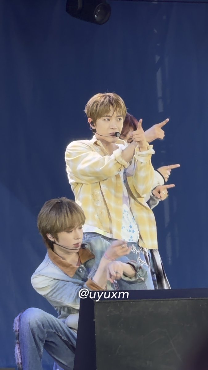 Two male performers on stage, one in a yellow patterned shirt pointing upward and the other in a denim jacket seated, with the watermark '@uyuxm'.