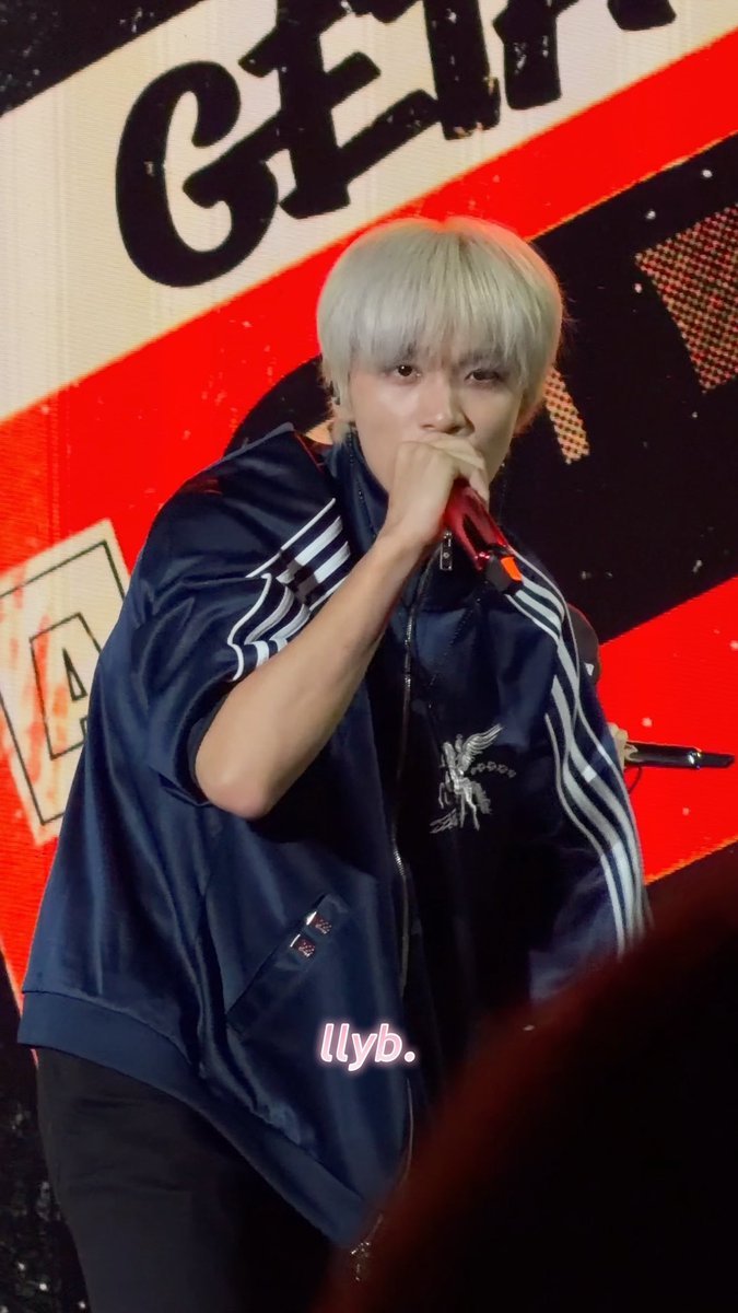 A performer with platinum blonde hair, wearing a navy Adidas jacket with an eagle logo, singing into a red microphone; image watermarked 'llyb.'