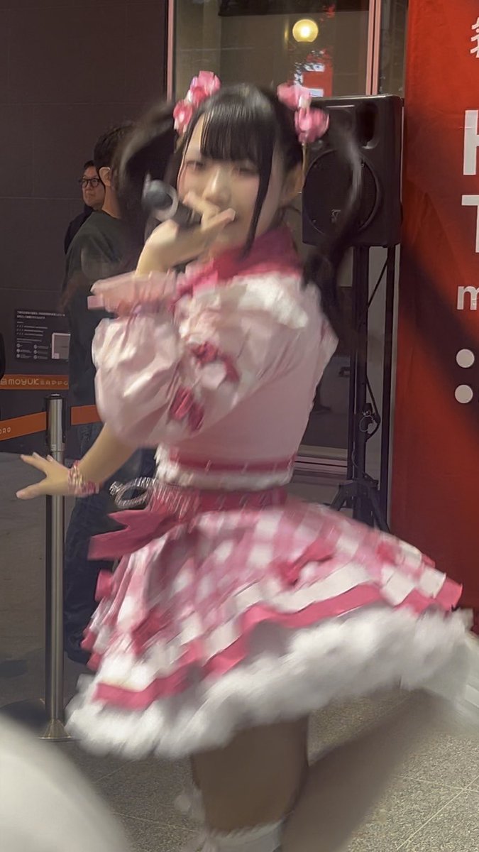 A young idol in a frilly pink and white checkered dress sings into a microphone at an indoor event, with red promotional banners visible in the background.