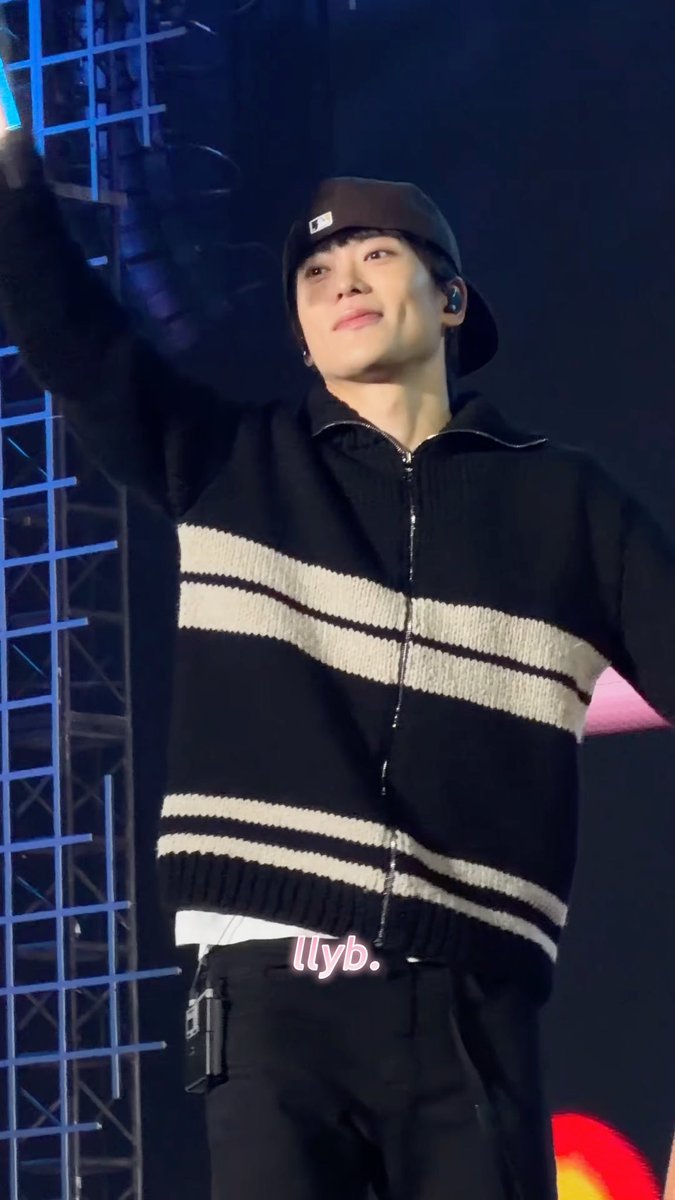 NCT127's Jaehyun waves to fans while wearing a black striped cardigan and backwards cap during the concert in Jakarta, with a pink "Ilyb." watermark visible on the image.