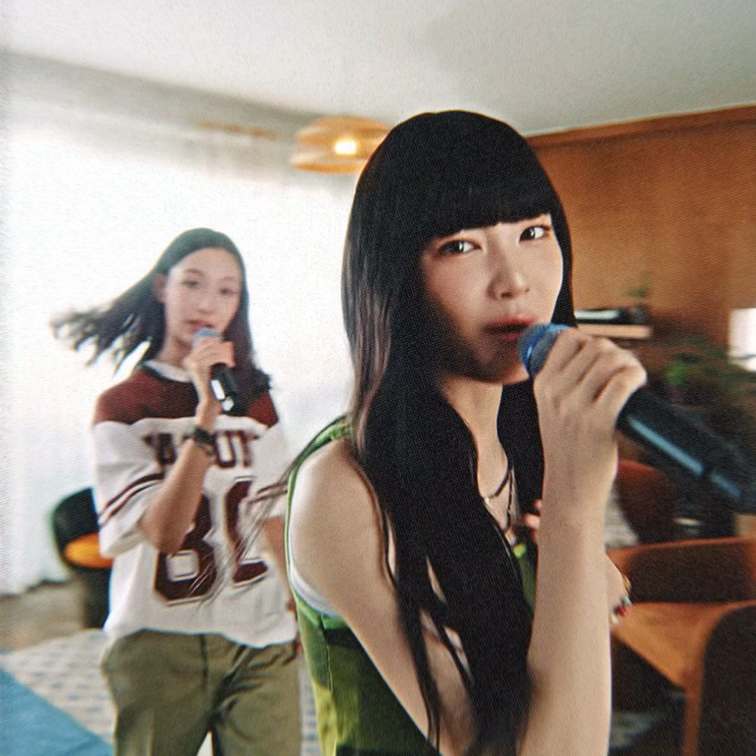 Two women are singing into microphones in an indoor setting, with the woman in the foreground wearing a green top and long black hair with bangs.