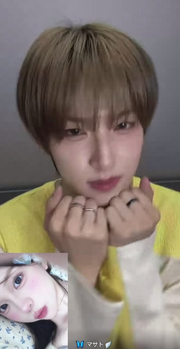 A selfie-style video call screenshot of a young man with brown hair wearing a yellow sweater, his hands clasped near his face, with a smaller inset video of a woman visible in the bottom left corner and a butterfly filter effect at the bottom.
