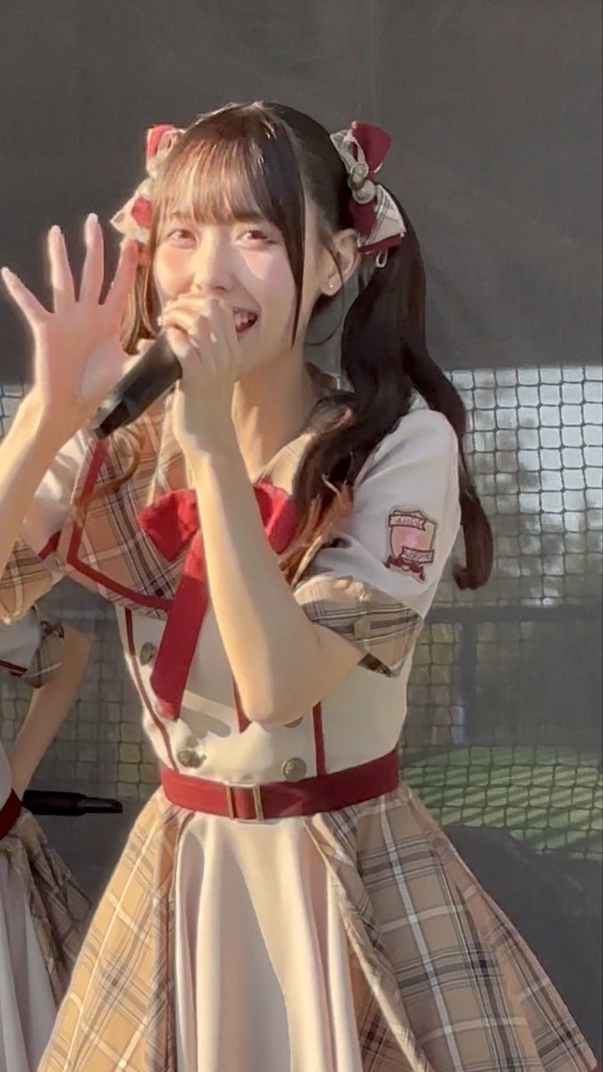 A young woman in a red and cream plaid idol costume sings into a microphone while waving, with pigtails tied by red ribbons.