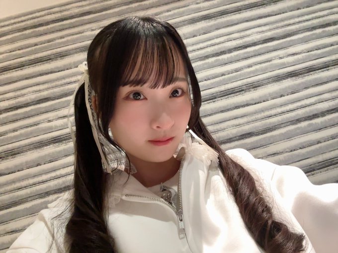 A young woman with long dark hair styled in pigtails with white lace ribbons poses for a selfie, wearing a white zip-up jacket against a striped background.