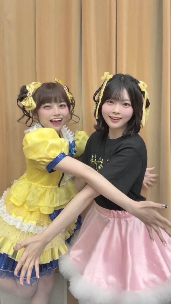 Two young women pose together indoors; one wears a yellow and blue frilly dress with yellow bows in her hair, while the other wears a black t-shirt with a pink skirt and yellow hair bows.