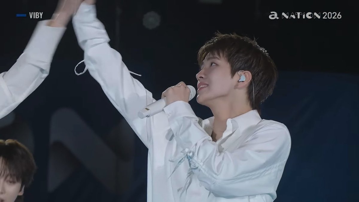 A young male performer in a white shirt sings into a microphone on stage with one arm raised, with "VIBY" and "a NATION 2026" logos visible.