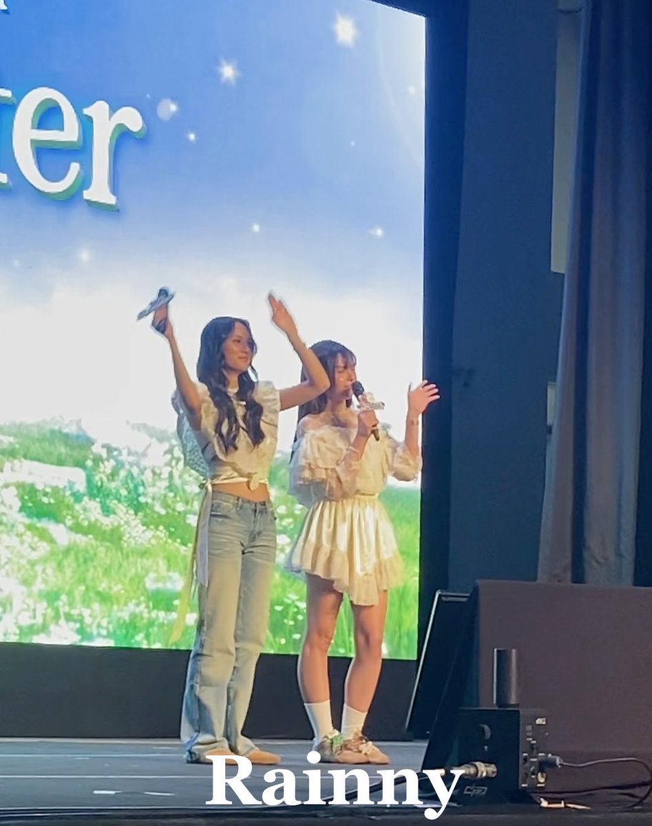Two young women on stage at what appears to be a fan event, one in jeans and a white top waving, the other in a cream dress holding a microphone, with 'Rainny' labeled at the bottom.