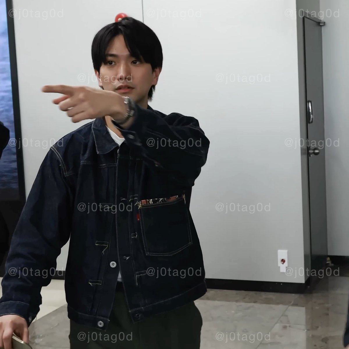 A young man with dark hair wearing a black denim jacket points toward the camera with a serious expression in what appears to be an indoor hallway setting. The image contains repeated "j0tag0d" watermarks across the entire frame, and the Japanese caption translates to criticisms about an unnecessary hand wave and a woman standing in front of someone named Kojiken, suggesting this is likely a screenshot from a behind-the-scenes video that became controversial.