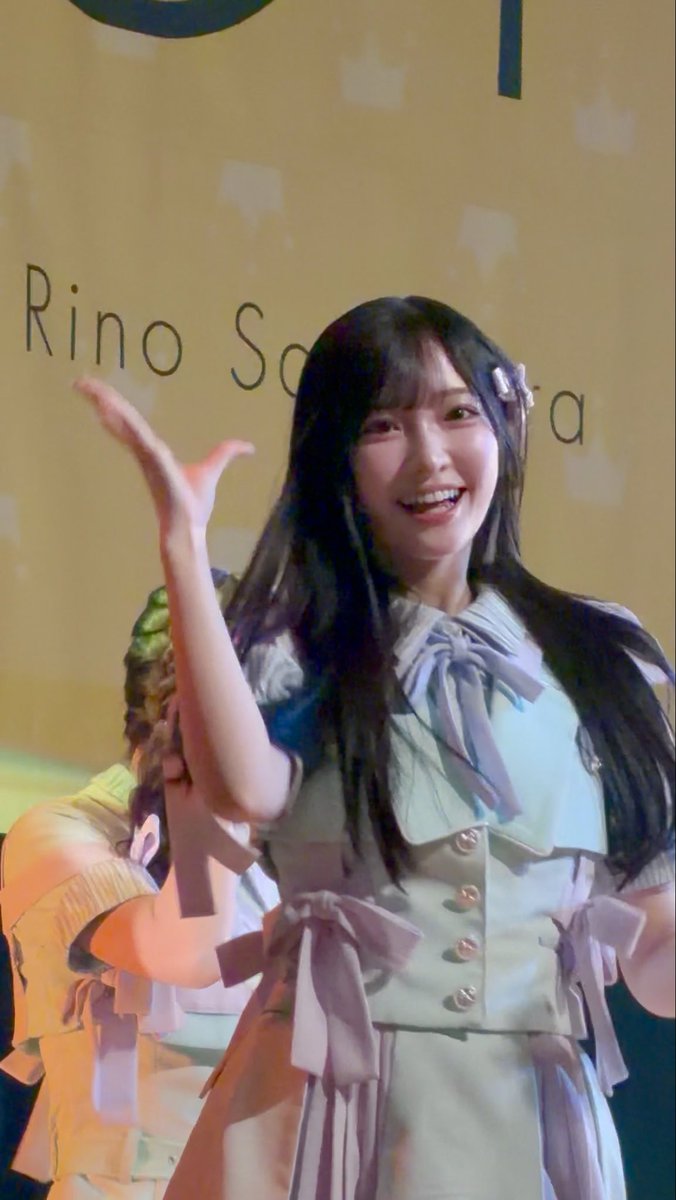 A smiling young woman with long dark hair waving her hand on stage in front of a backdrop that reads 'Rino S', wearing a light blue outfit with bow details.