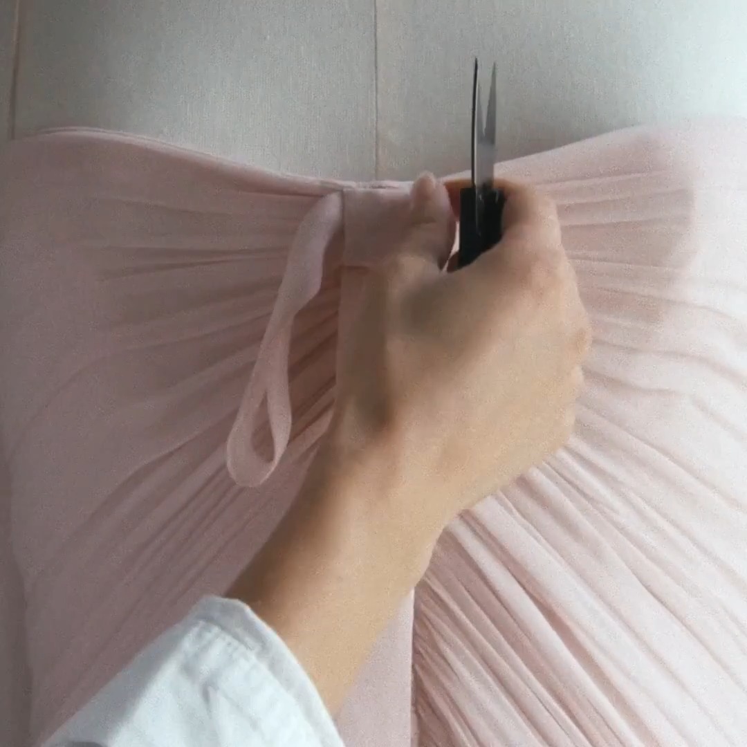 A close-up shows a hand using scissors to cut a ribbon on a pleated pastel pink chiffon garment, evoking the delicate craftsmanship behind Dior Summer 2027's revival of the 1957 Zerline dress.