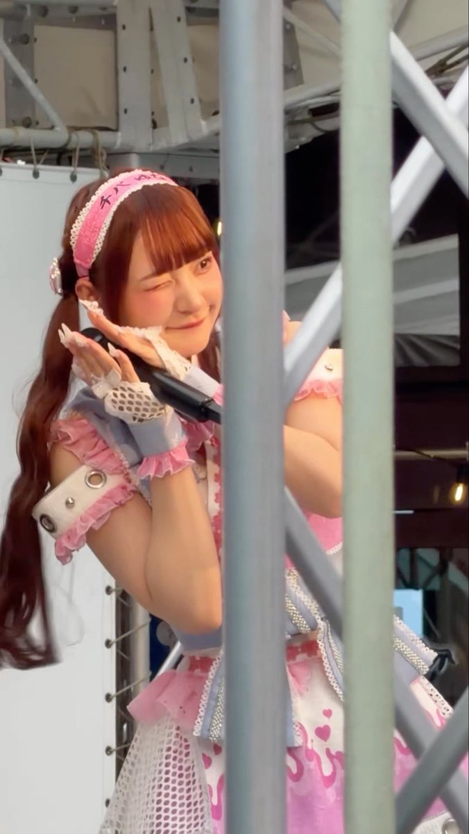 A young woman in a pink and white frilly idol costume with a matching headband winks and poses playfully behind metal scaffolding at what appears to be a stage event.