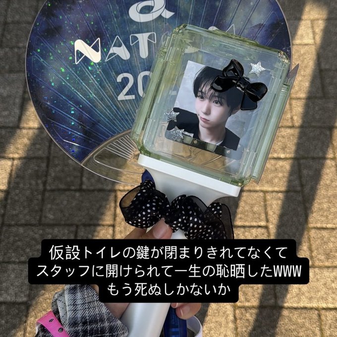 A hand holds a K-pop lightstick displaying a member's photo with a black bow, with Japanese text overlay about being embarrassed when staff had to open a bathroom door.