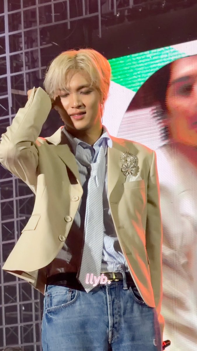 A young man with blonde hair performs on stage wearing a beige blazer with a decorative brooch, a blue striped shirt, and a loose tie, with the watermark "llyb." overlaid.