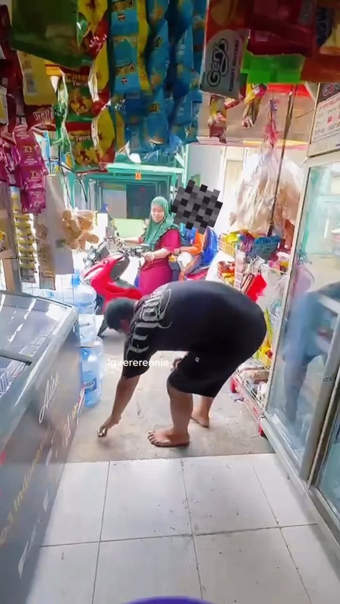 A barefoot man bends down to pick up scattered money on the floor of a small convenience store, while a woman sits on a red motorbike near bottled water (Aqua) and snacks. The scene illustrates the post's complaint about someone rudely tossing 100k Indonesian rupiah onto the ground after buying only two bottles of water.