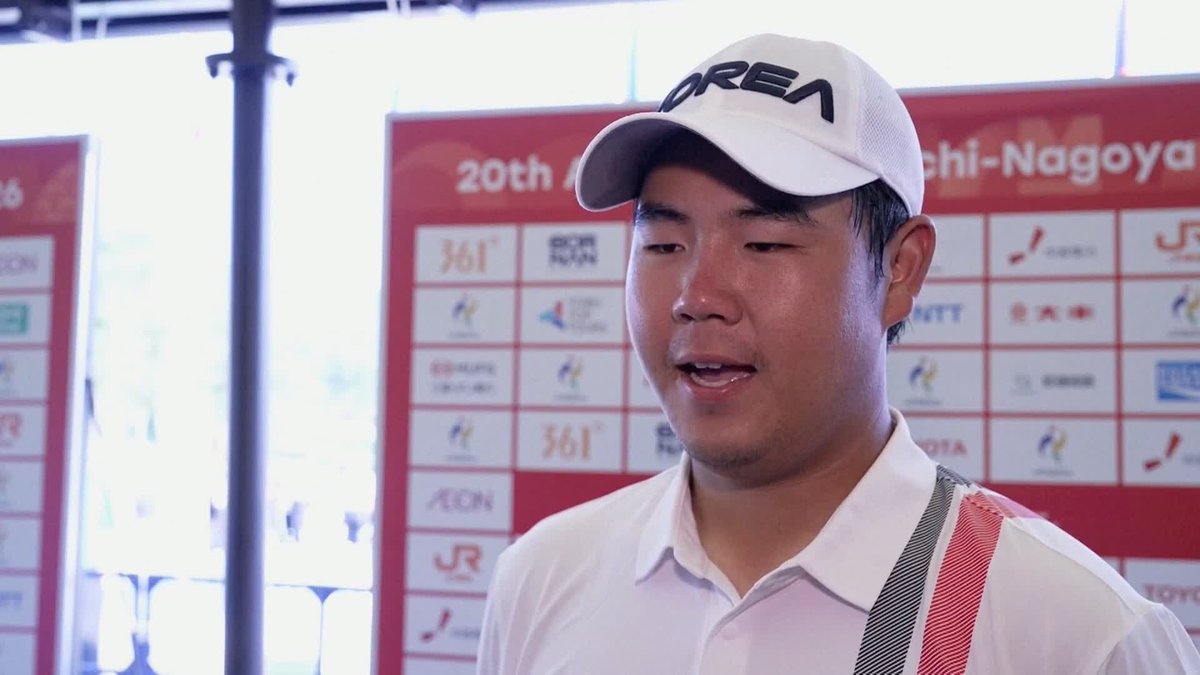A young man wearing a white cap appears to be speaking during an interview at what seems to be a sporting event, with a backdrop showing sponsor logos like "361°," "AEON," "JR," and text reading "20th... Nagoya." The image likely depicts Tom Kim, the South Korean golfer who secured an exemption from full military service after winning gold at the Asian Games, as referenced in the caption.