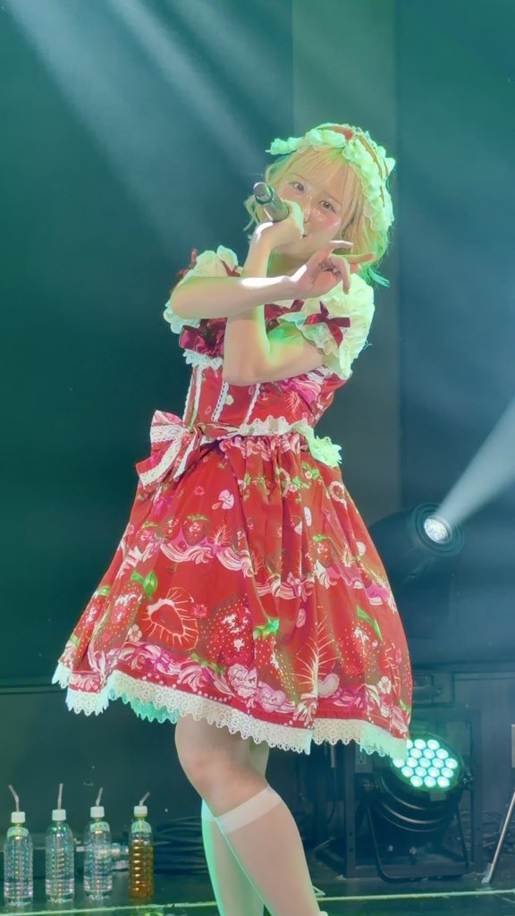 A performer with blonde hair in a strawberry-patterned red dress sings into a microphone on stage under green stage lights.