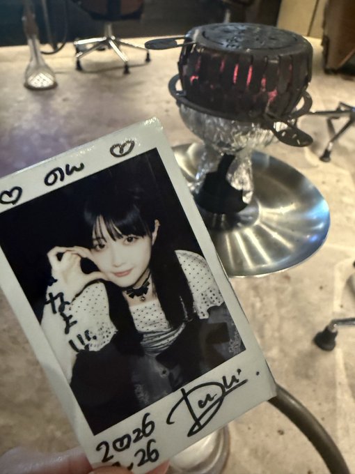 A black and white signed polaroid photo of a young woman making a heart shape with her hands, dated '2026.6.26', held up in front of a small tabletop grill.