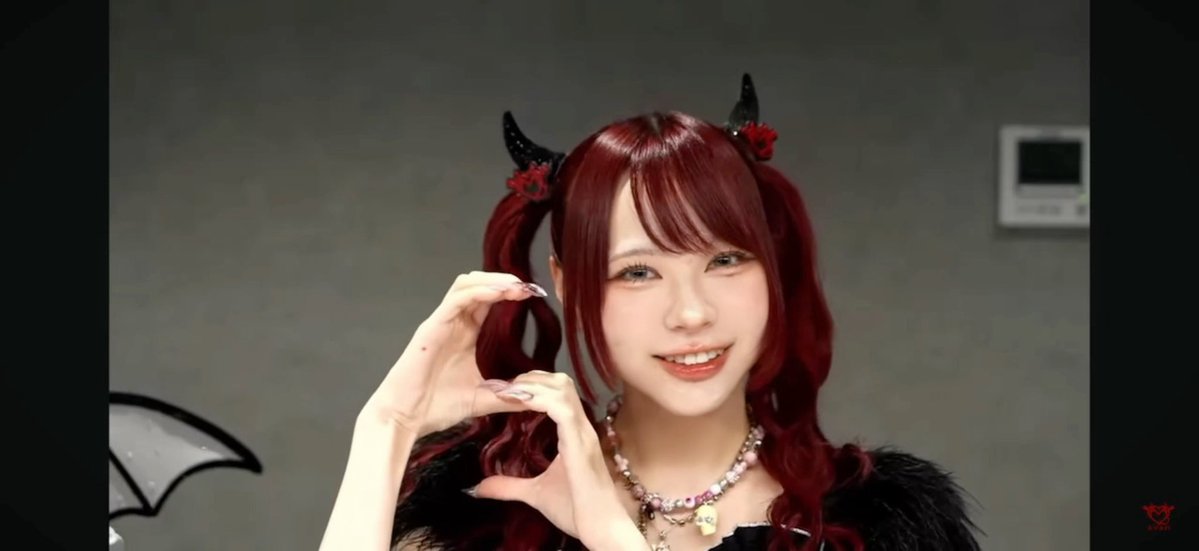 A cosplayer with dark red hair styled in twin tails with black horns poses making a heart shape with her fingers, wearing a furry black outfit.