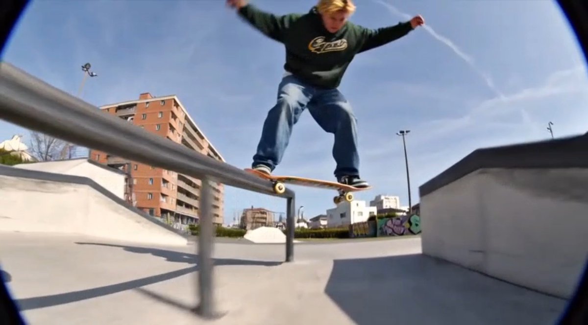 An action shot of a skateboarder in a green hoodie and blue jeans performing a trick, grinding along a rail at a concrete skate park.