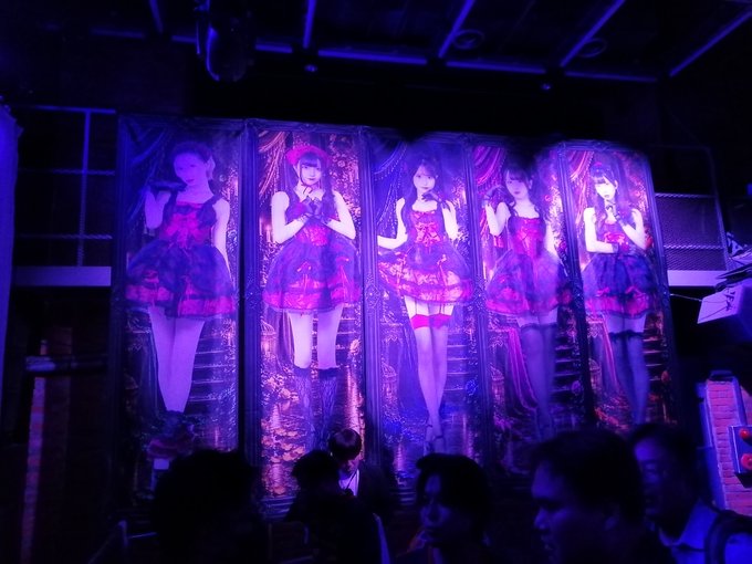 Female idol performers in matching pink dresses are projected onto large transparent screens at a live concert venue bathed in purple lighting.