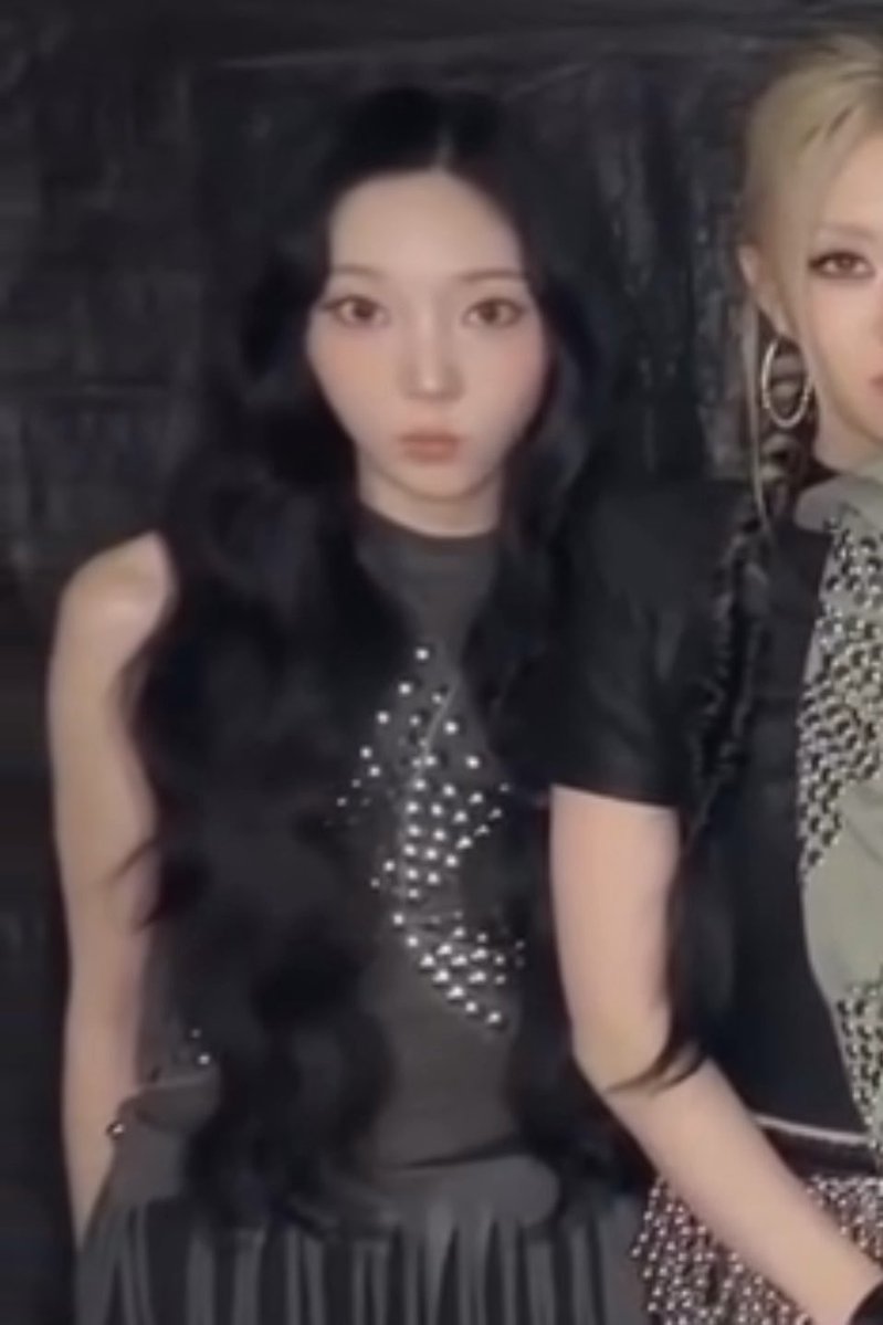 Two women pose together in dark outfits, with the central figure wearing a black top adorned with pearl embellishments and long wavy black hair.