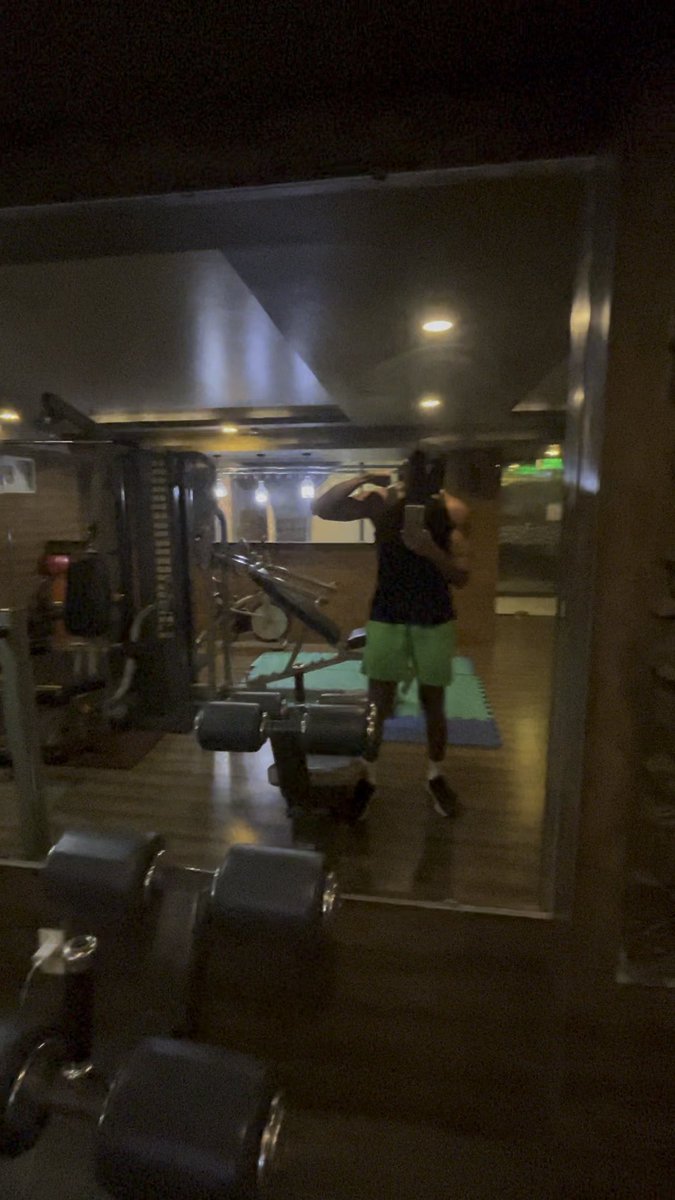 A blurry mirror selfie of a person in a black tank top and green shorts posing with a flexed arm in a dimly-lit home gym setting.