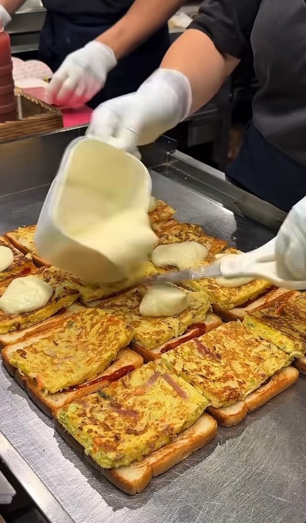 Gloved hands pour melted cheese from a container onto a tray of egg-and-bread sandwiches during food preparation.