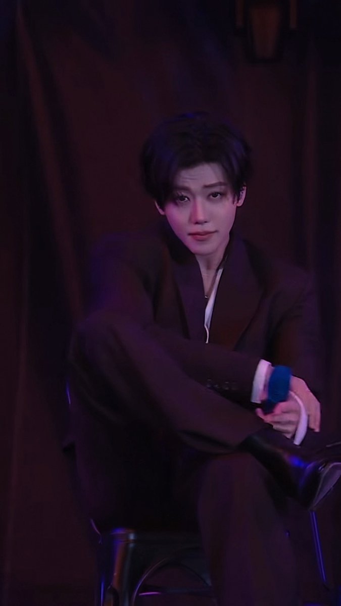 A young man with dark hair sits in a dimly lit setting with purple-toned lighting, wearing a black suit and holding what appears to be a small object, gazing directly at the camera with a soft, contemplative expression.

The post in Thai translates roughly to "May I bow to you, older brother?" — a phrase that complements the image's gentle, almost reverent mood, as the man's serene posture and direct gaze seem to welcome such a humble request.