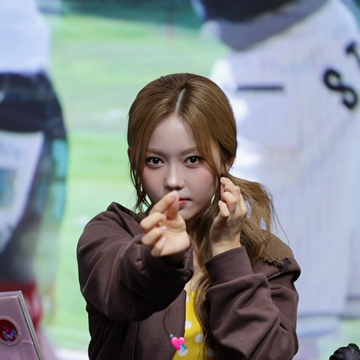Portrait photo of a young woman with light brown hair in a side ponytail, wearing a brown zip-up hoodie over a yellow polka-dot top, pointing toward the camera with a small pink heart sticker overlaid on the image.