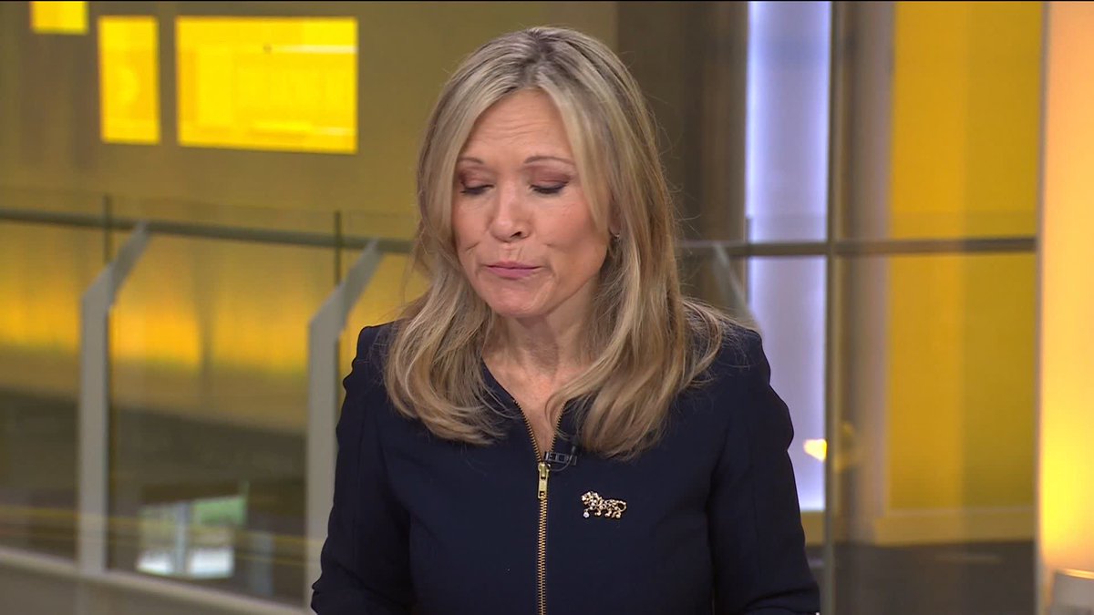 A blonde female news anchor wearing a dark navy jacket with a leopard brooch is shown with her eyes closed or looking down, appearing emotional, in a news studio with yellow-lit background.