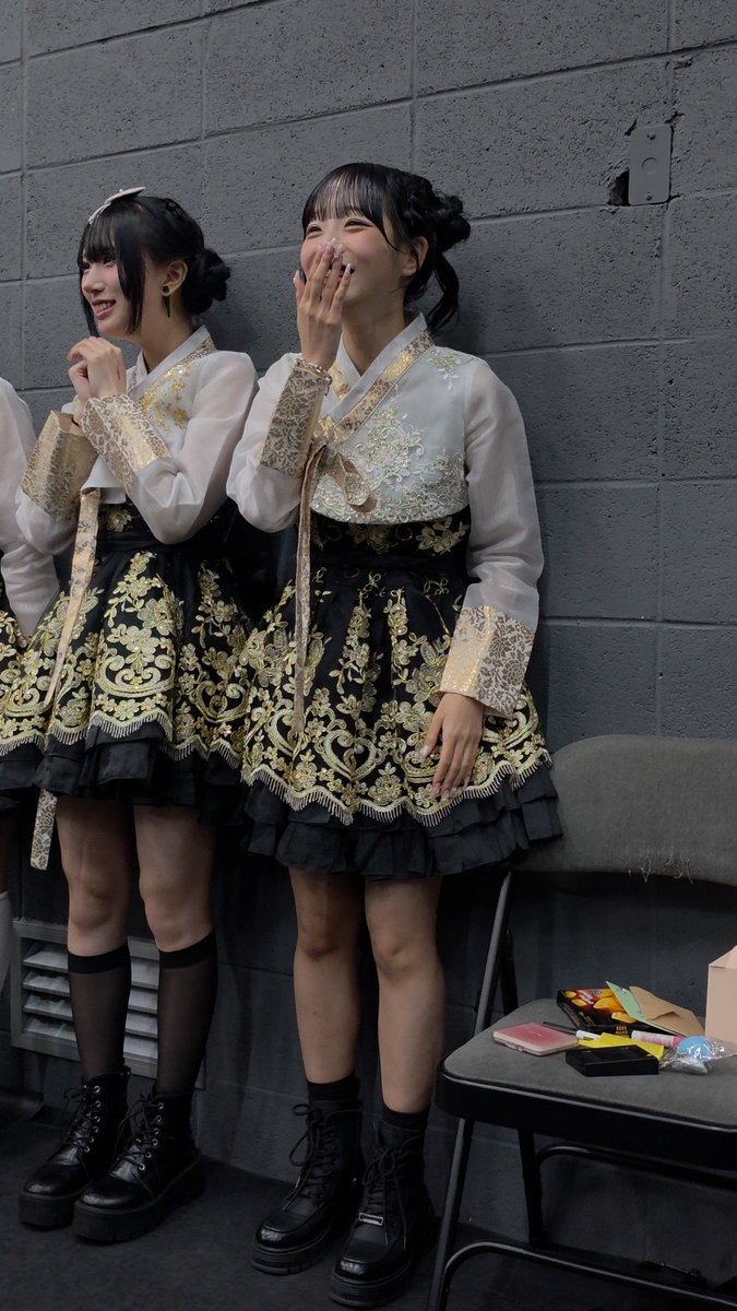 Two young women wearing matching ornate black and gold hanbok-style costumes and platform boots laugh backstage, leaning against a gray brick wall.