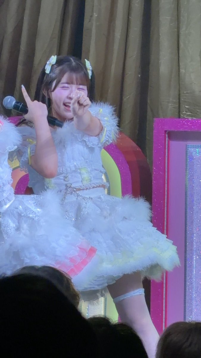 A performer in a fluffy light blue idol dress winks and points at the camera while holding a microphone on a stage with colorful decorations.