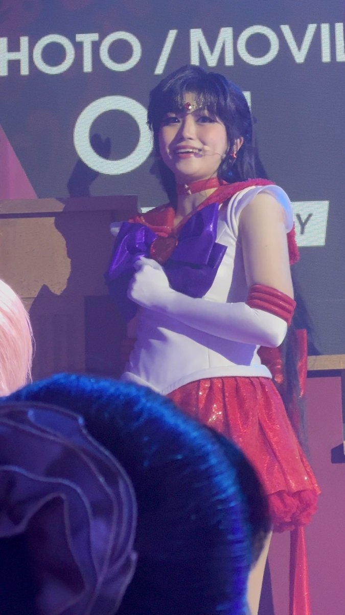 A performer in a Sailor Moon-style sailor suit costume with a white top, red skirt, and long gloves performs on stage in front of a screen reading 'HOTO / MOVI'.