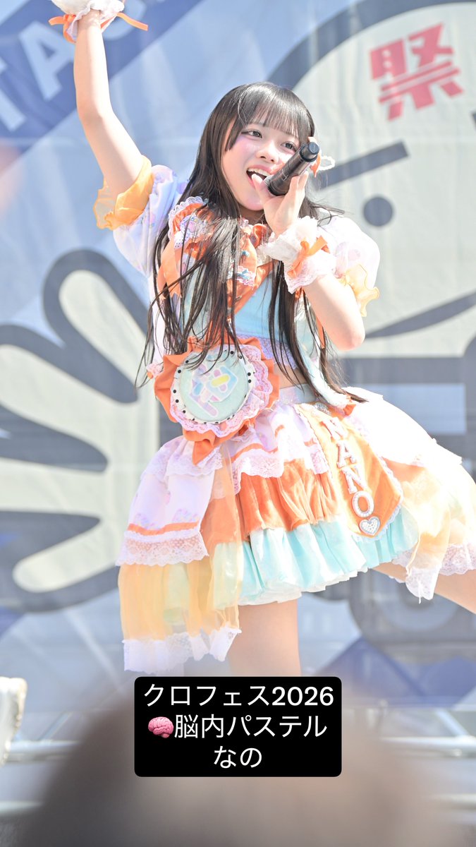 An idol performer on stage wearing a colorful orange, white, and turquoise frilly outfit, singing into a microphone with one arm raised, with a text overlay reading "クロフェス2026 🧠 脳内パステル なの".