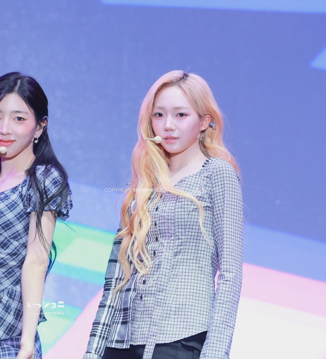 Two performers on stage; the woman in focus has long blonde hair and wears a plaid gray outfit with an earpiece microphone, while another performer in a blue plaid top stands to the left.
