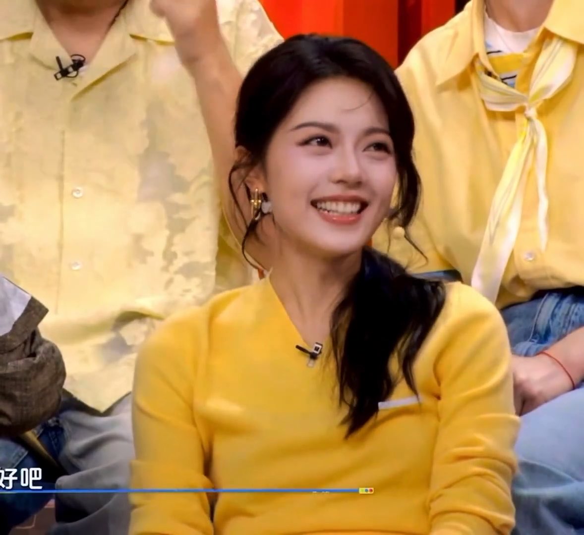 A young woman with dark hair wearing a yellow sweater smiles brightly on what appears to be a Chinese television program, with Chinese text visible at the bottom left.