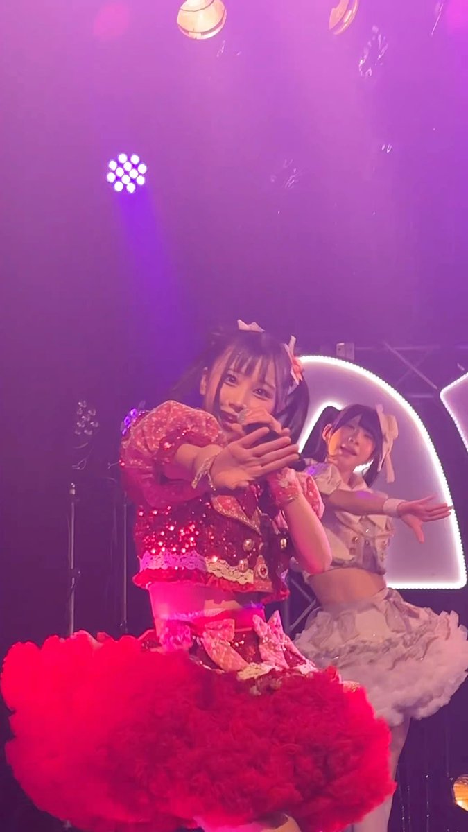 A female performer in a sparkly red idol dress is singing into a microphone on a purple-lit stage, with another performer in a white outfit visible behind her.