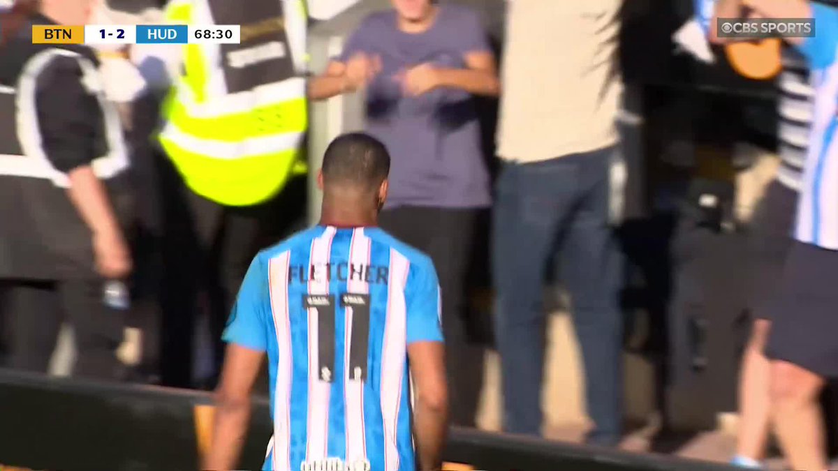 Huddersfield Town player Fletcher #11 walks near the crowd during a match against BTN shown at 68:30, with the score 1-2 in favor of HUD. The image provides visual context to the commentary discussing goalkeeper Corey Addai's positioning on a goal-line incident.