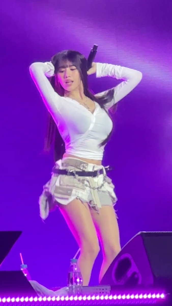 A female performer on stage with a purple backdrop dances energetically, wearing a tight white long-sleeved top and a layered denim skirt with chains.