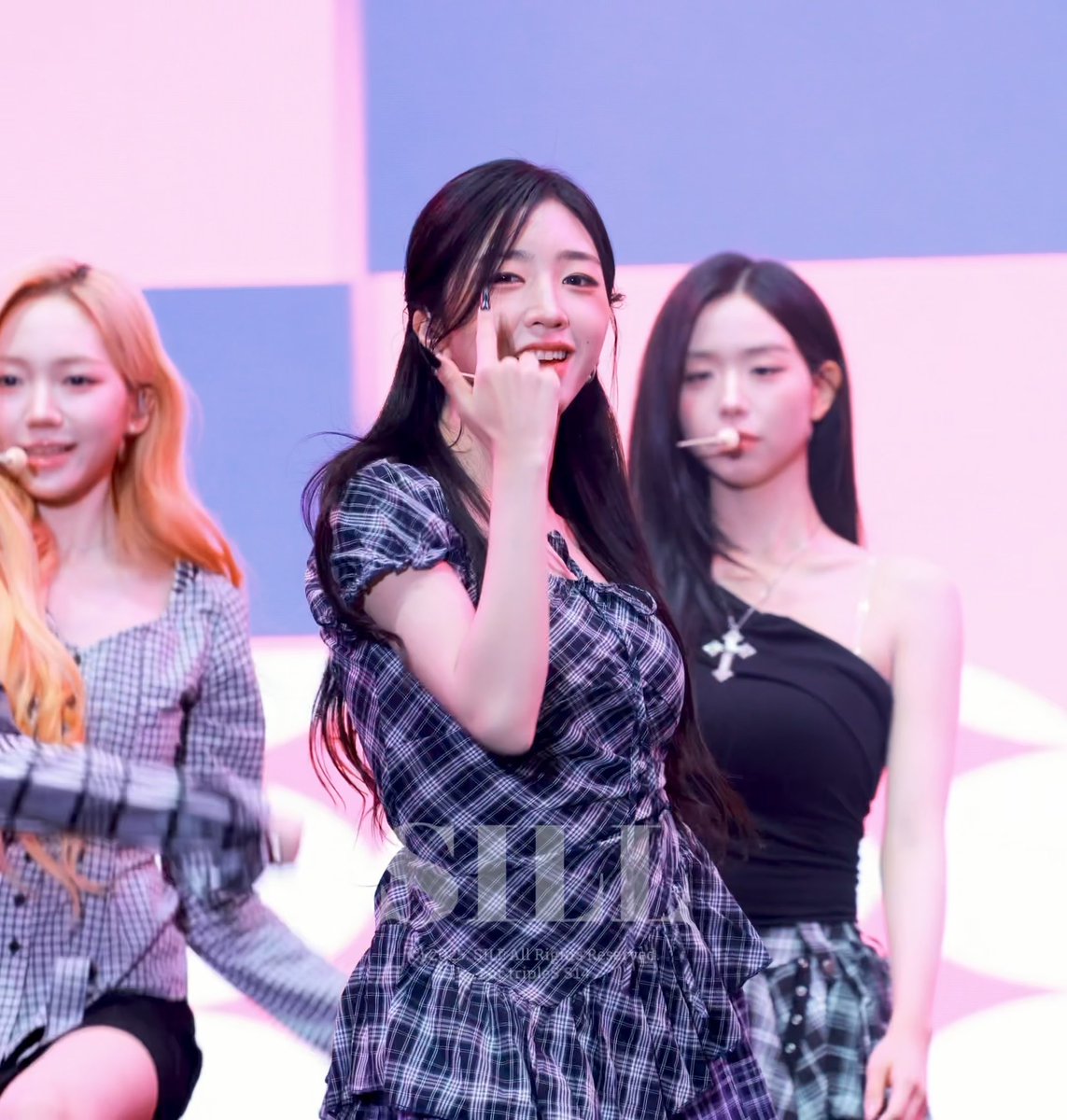 Three women performing on stage; the center woman in a plaid dress smiles and makes a finger heart gesture, while the others wear plaid and black outfits.