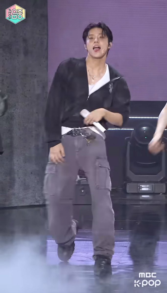A male K-pop performer in a black zip-up jacket, white undershirt, and gray cargo pants performs on stage, with 'MBC K-POP' logo visible in the corner.