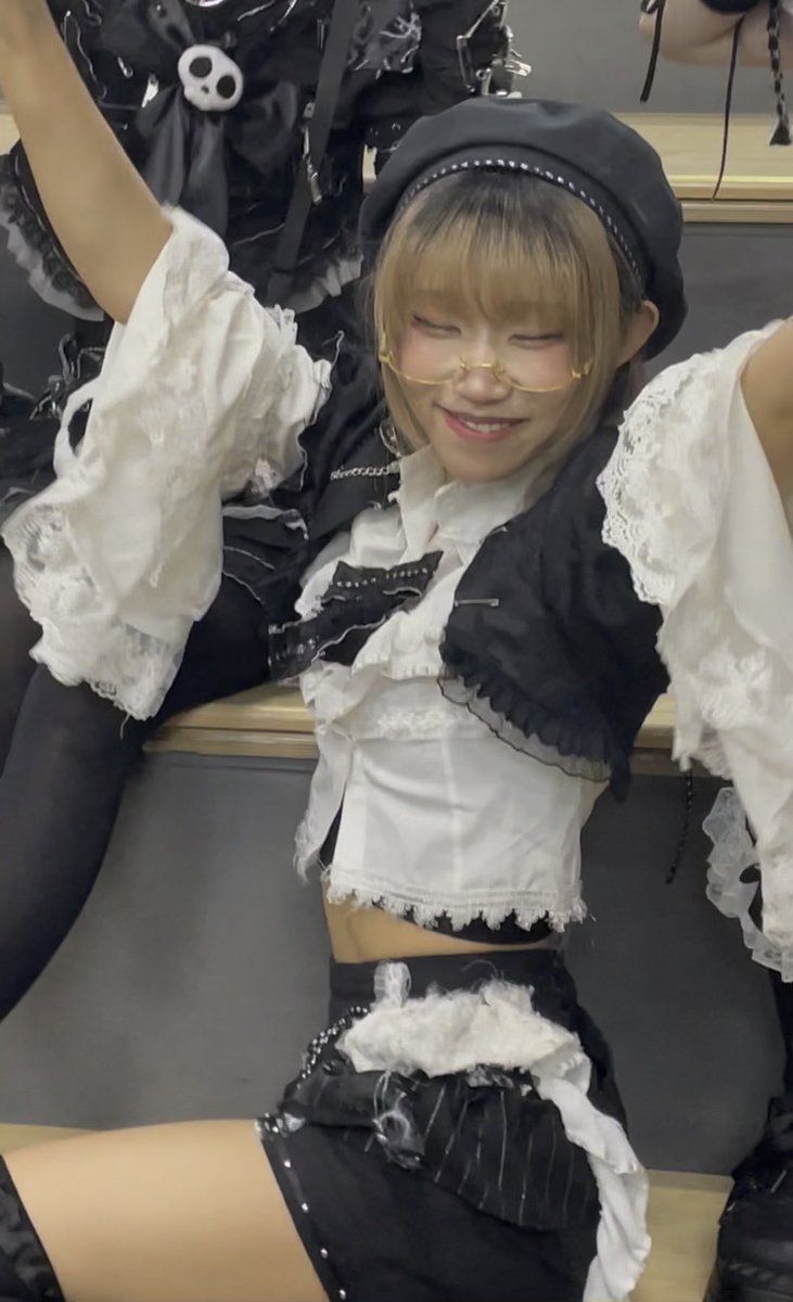 A young woman with blonde hair, glasses, and a black beret, wearing a black and white frilly outfit, smiling with her eyes closed and arms raised in what appears to be a maid-style costume.