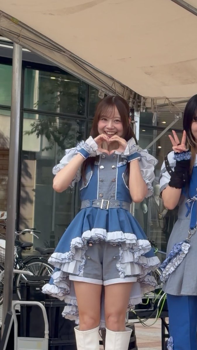 A smiling young woman in a blue and white ruffled idol dress with matching shorts, making a heart shape with her hands.