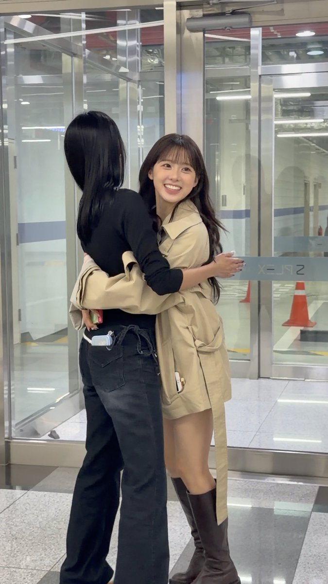 Two women are warmly hugging in what appears to be an airport or transit terminal, with one wearing a beige trench coat and brown boots, and the other in a black top and dark jeans.
