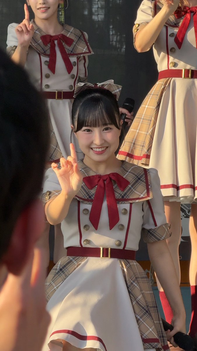 A young woman with bangs smiles brightly while making a peace sign, wearing a cream and plaid school uniform-style idol outfit with red accents, with other similarly dressed performers visible behind her.