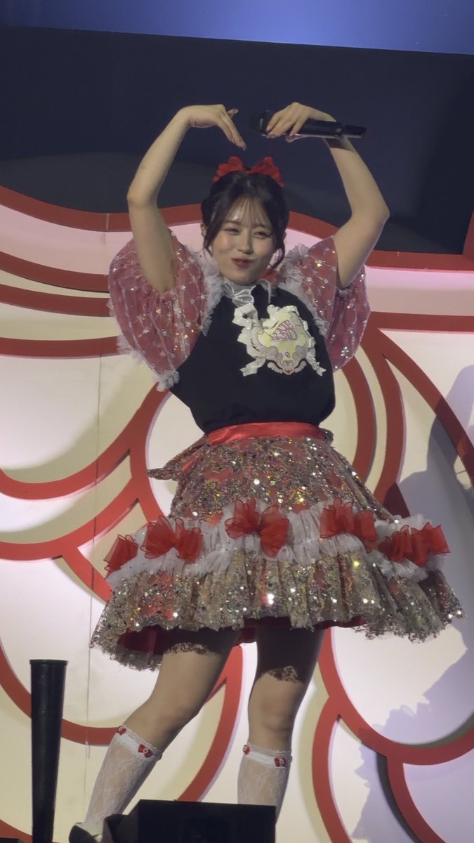 A performer in a black, pink, and white frilly costume with a red bow strikes a pose holding a microphone above her head, with a red and white swirl backdrop behind her.